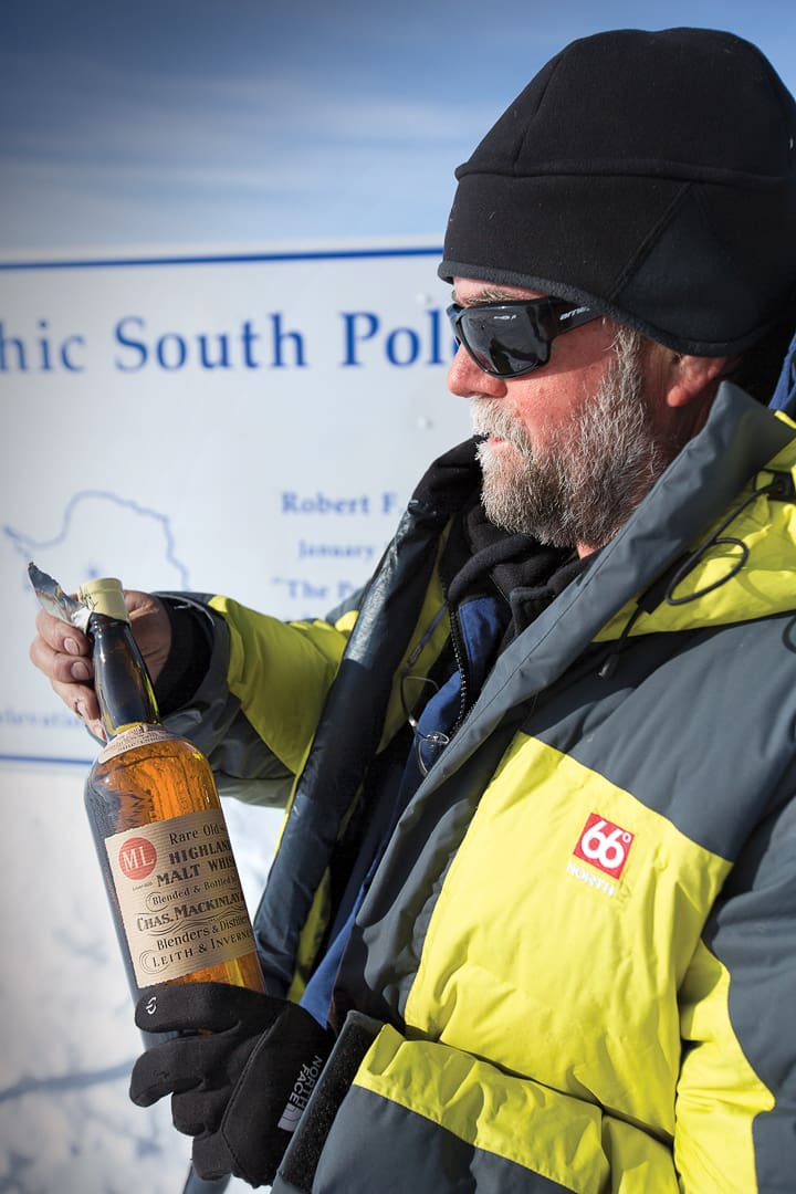 A person in a yellow and grey parka, hat, and sunglasses holds a bottle of Mackinlays Rare Old Highland Malt Whisky in front of the South Pole sign during an Expeditions 7 journey.