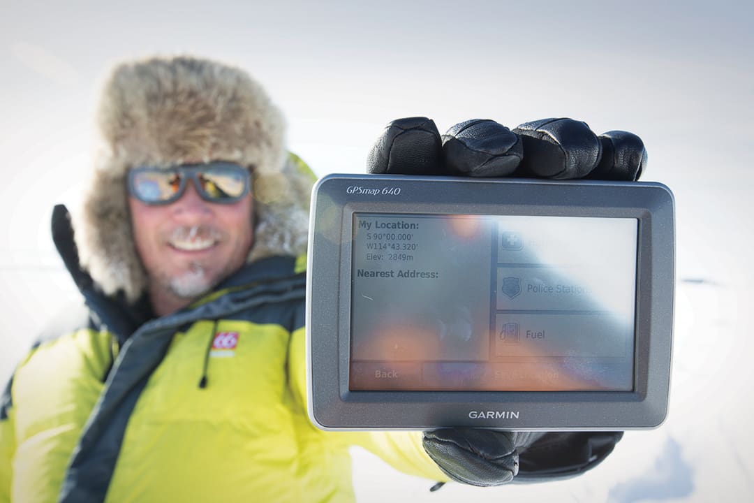 A person wearing winter gear holds up a Garmin GPS device displaying coordinates and location icons during an Expeditions 7 trek across the White Desert.