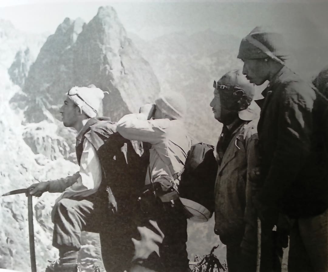 Four mountaineers wearing climbing gear look out over rugged, rocky peaks, appearing focused and determined. The black-and-white photo evokes the adventurous spirit of the Omidvar brothers’ historic journeys.