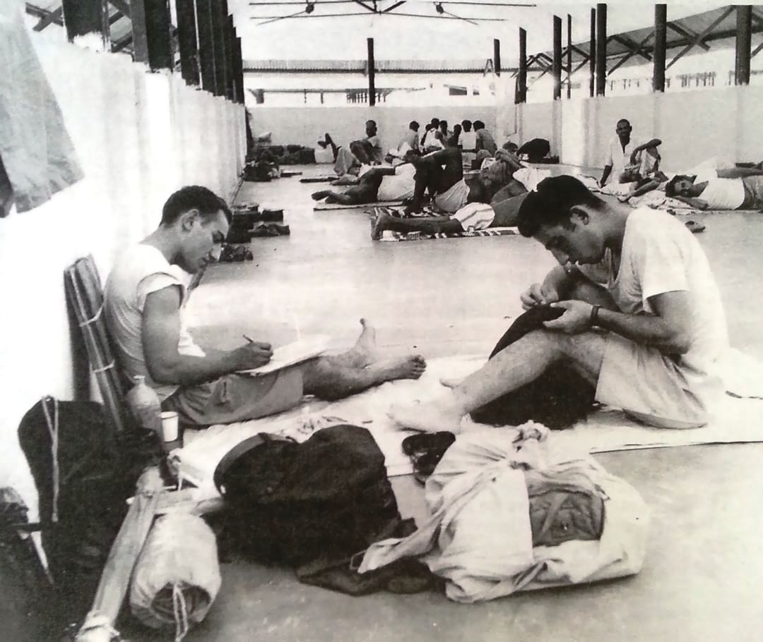 Men sit and lie on thin mats in a large, open shelter, some writing or reading. Belongings are scattered around them, evoking scenes from the Omidvar brothers’ adventurous communal travels.