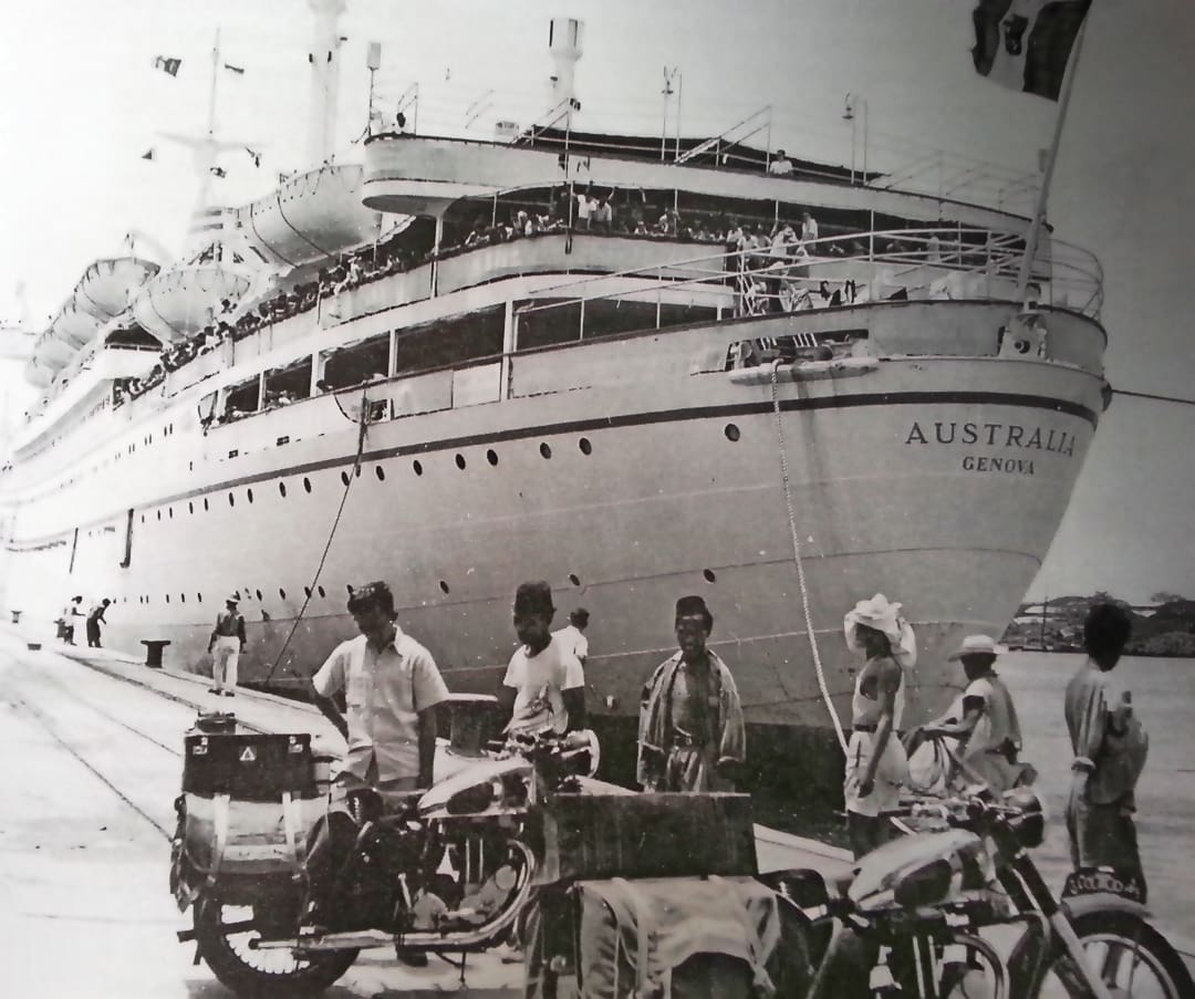A large passenger ship named Australia Genova is docked. The Omidvar brothers stand with others on the pier, surrounded by bicycles, motorcycles, and luggage in this black-and-white photo. Some look at the camera.