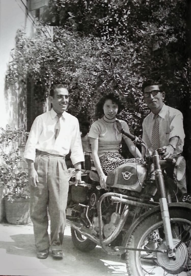 Three people pose next to a motorcycle; a woman sits on the bike while two men, resembling the Omidvar brothers, stand beside her in mid-20th-century attire. Bushes and a building are in the background.