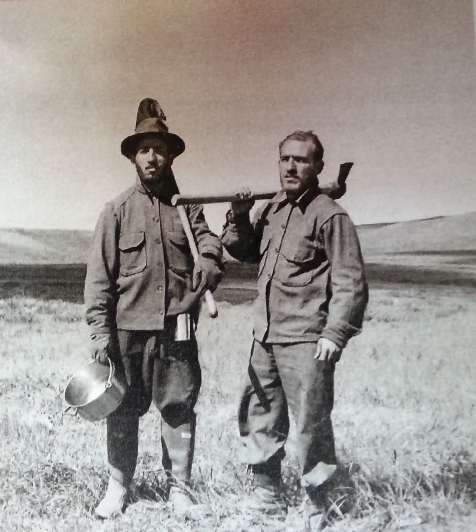 Two men in work clothes stand in a grassy field. One holds a hatchet on his shoulder, the other a cooking pot. Hills stretch out behind them. Black and white photo, possibly showing the Omidvar brothers in a historical setting.