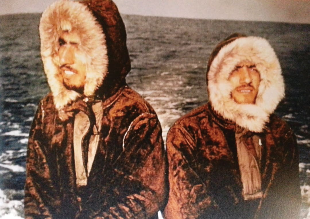 Two people wearing fur-lined hooded parkas stand side by side near a body of water, reminiscent of the Omidvar brothers, with the ocean visible in the background.