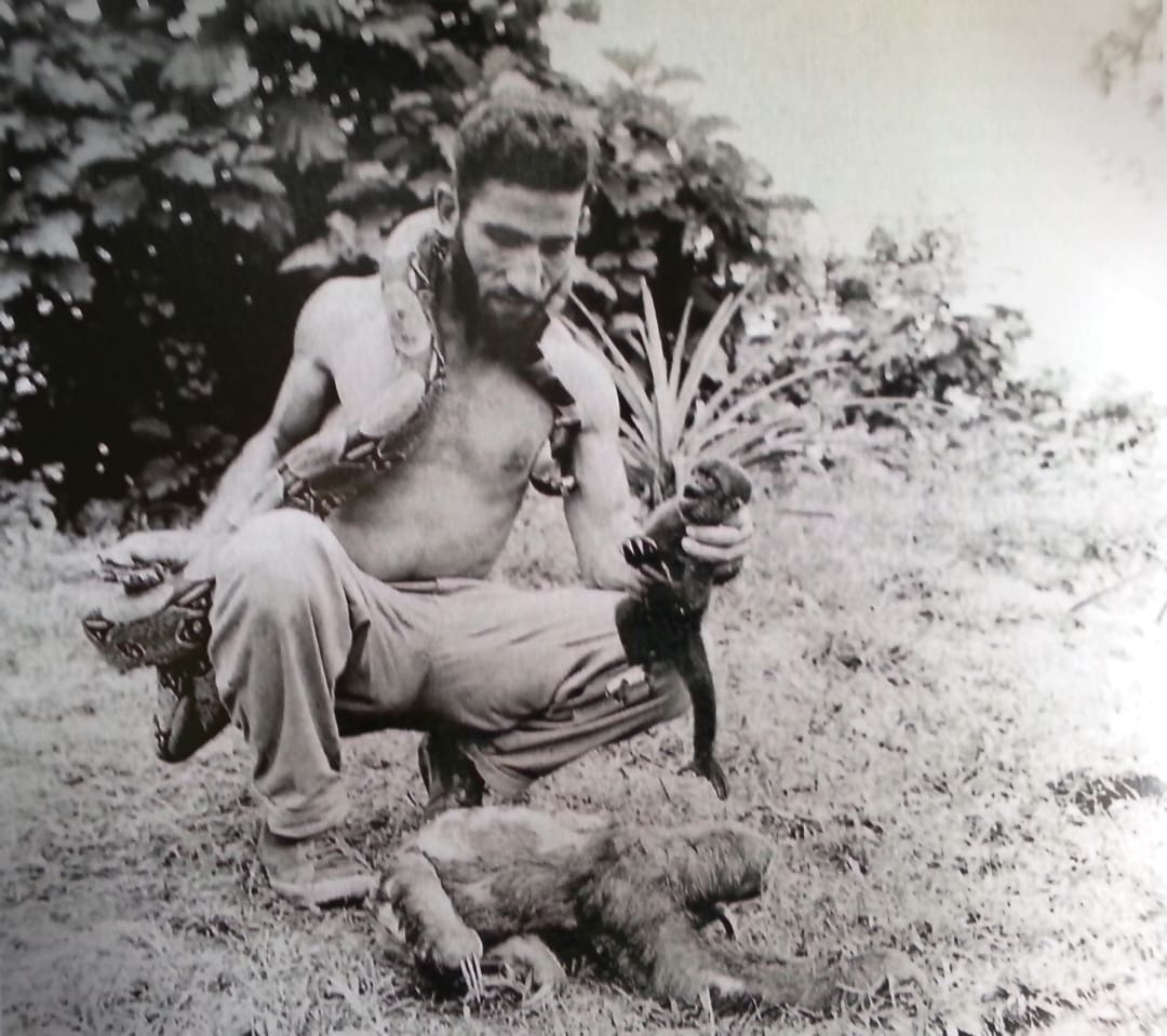A shirtless man, reminiscent of the adventurous Omidvar brothers, crouches outdoors holding a snake around his shoulders, an otter in one hand, and a sloth at his feet amidst lush foliage.