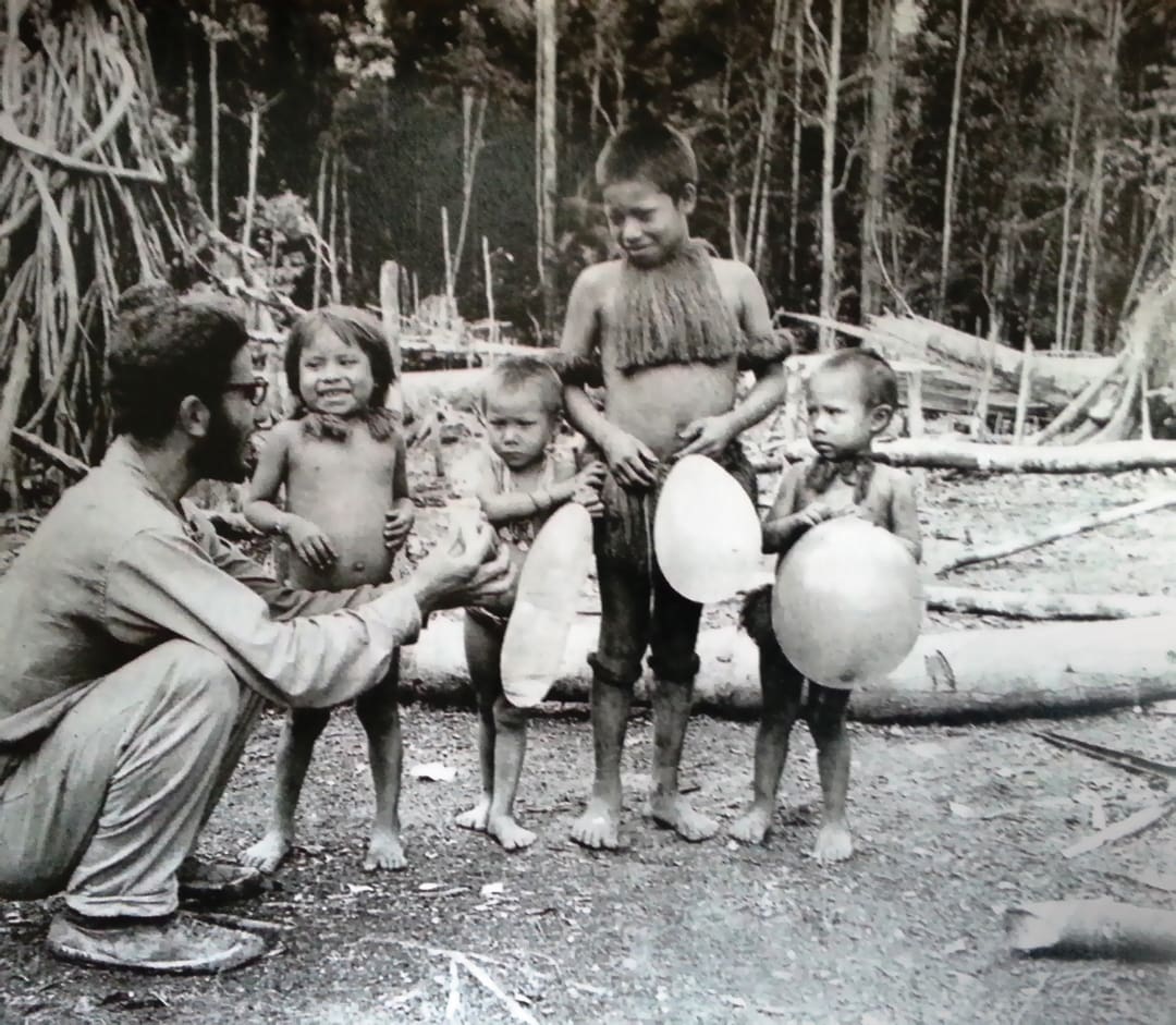 A man kneels and interacts with four young children holding balloons in a forest clearing, reminiscent of scenes photographed by the Omidvar brothers. The barefoot children wear minimal clothing, surrounded by trees and logs.