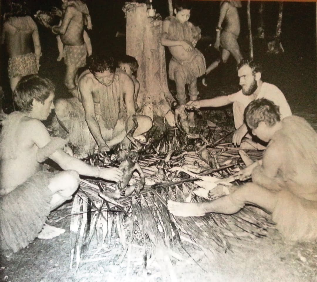 A group of people in primitive costumes, reminiscent of the Omidvar brothers’ explorations, sit in a circle around a fire preparing food, while others gather in the black-and-white background.