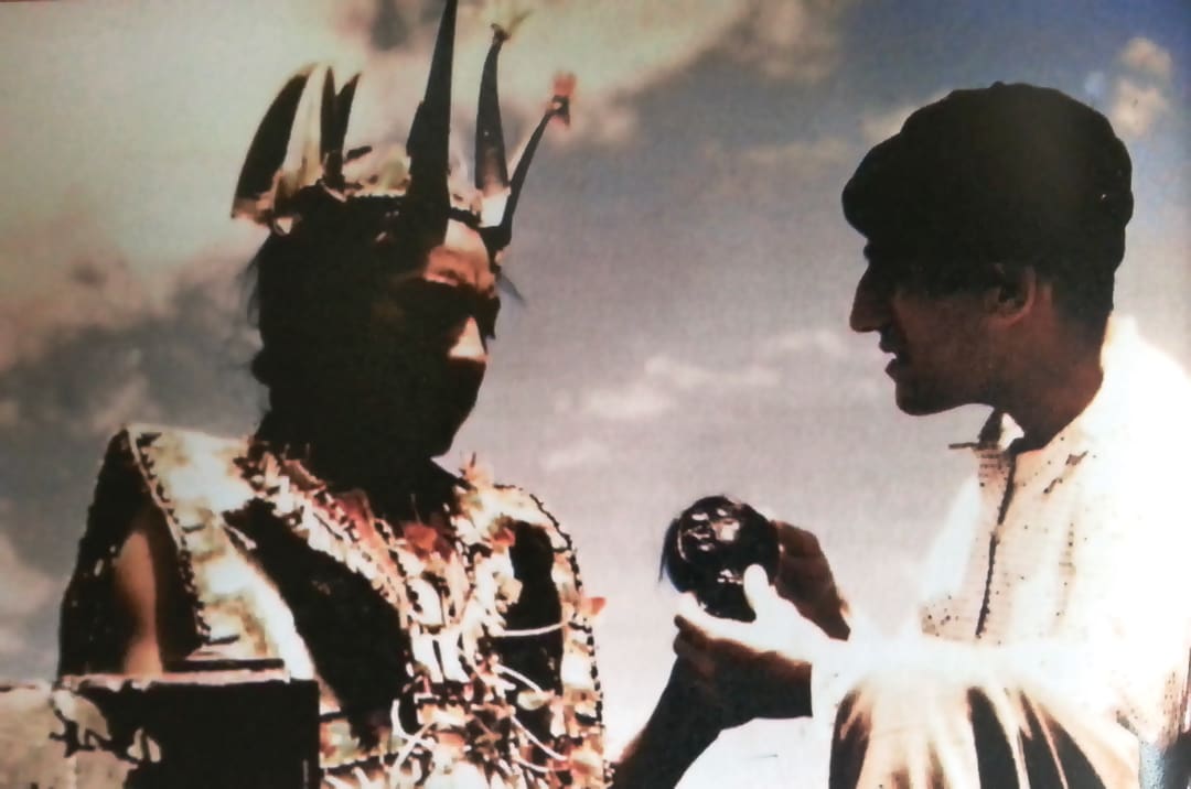 A person in elaborate traditional attire with a feathered headdress faces another, holding a dark round object—an encounter reminiscent of the Omidvar brothers’ adventurous journeys under a bright, cloudy sky.