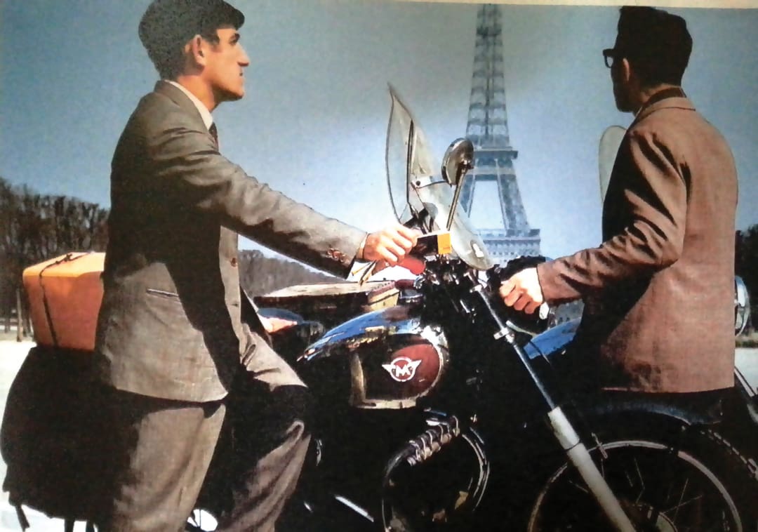 Two men in suits, reminiscent of the Omidvar brothers, stand beside a motorcycle with luggage near the Eiffel Tower in Paris, France, on a sunny day.