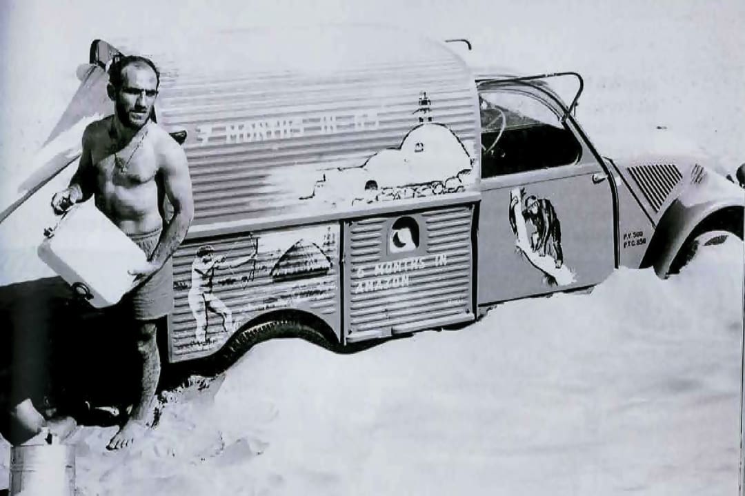 A shirtless man stands beside a partially buried car in the sand, holding a large white container. The car, reminiscent of the Omidvar brothers’ travels, has drawings of mountains and camels on its side in a desert setting.