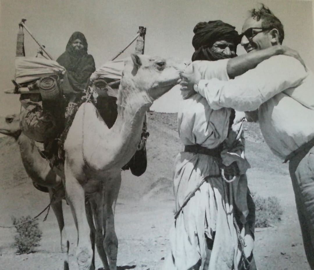 Two men, one in traditional clothing and one in modern attire—like the Omidvar brothers—greet each other warmly by a camel, where a woman sits among bundles in the desert landscape.