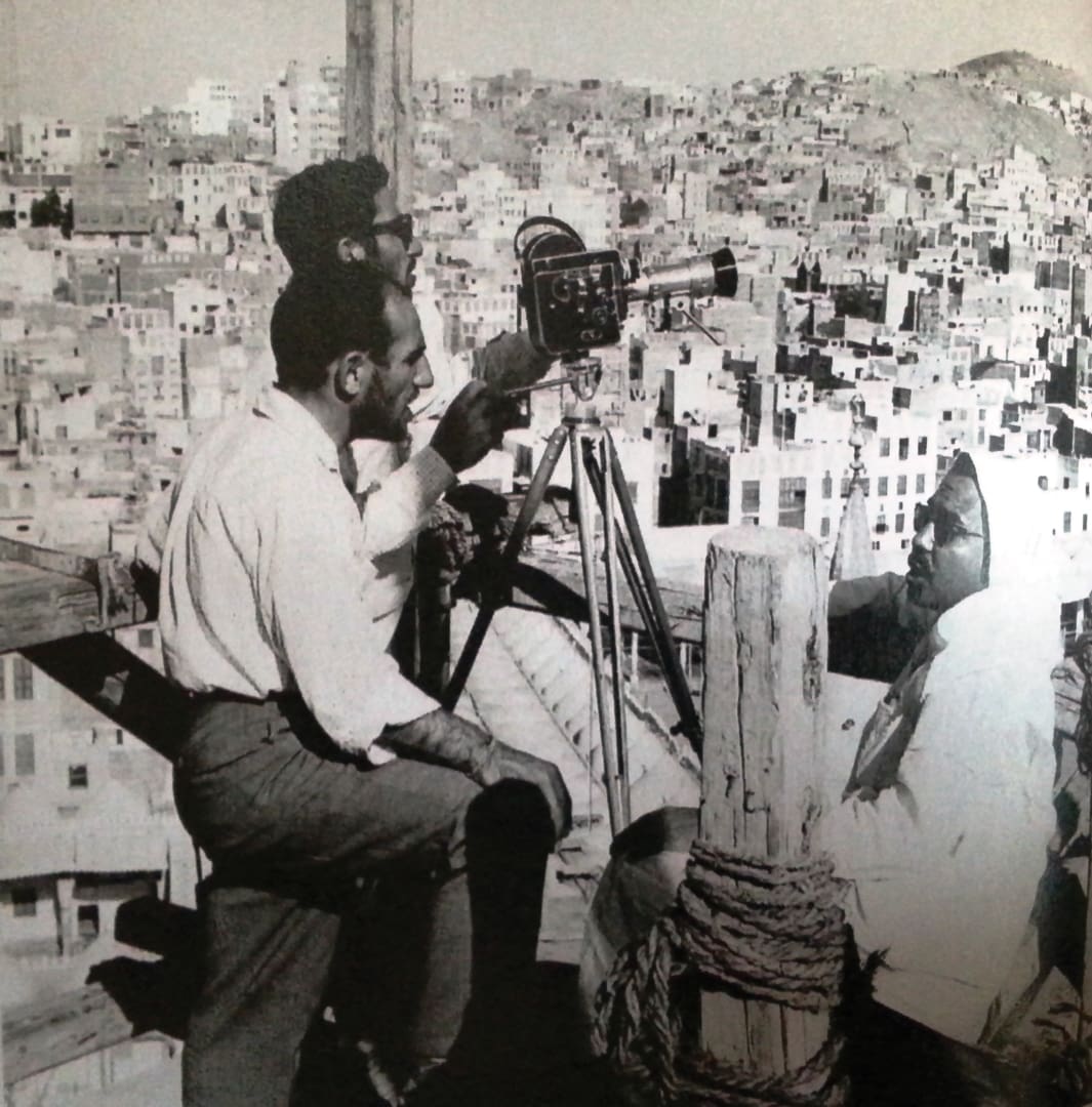 Two men, possibly the Omidvar brothers, operate a film camera on a tripod while filming a seated person outdoors, with a densely built hillside city in the background. The image is in black and white.