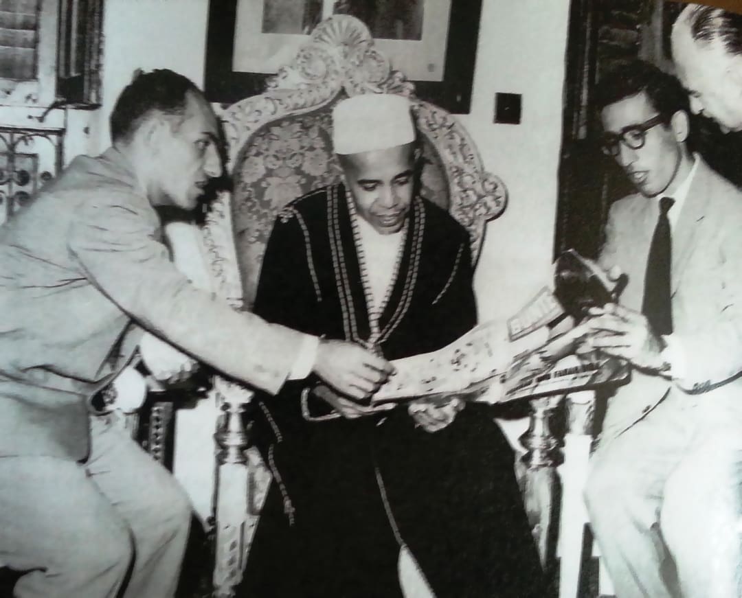 Four men sit together, two in suits and one in traditional attire with a fez, examining a newspaper—reminiscent of the Omidvar brothers engaged in thoughtful discussion in a formal, indoor setting.