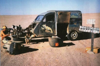 A person repairs a stripped-down car in a barren desert, reminiscent of the Omidvar brothers’ adventures. Animal artwork decorates the side, tires are scattered, and a sign reads SCIENCE ET MECHANIQUE.