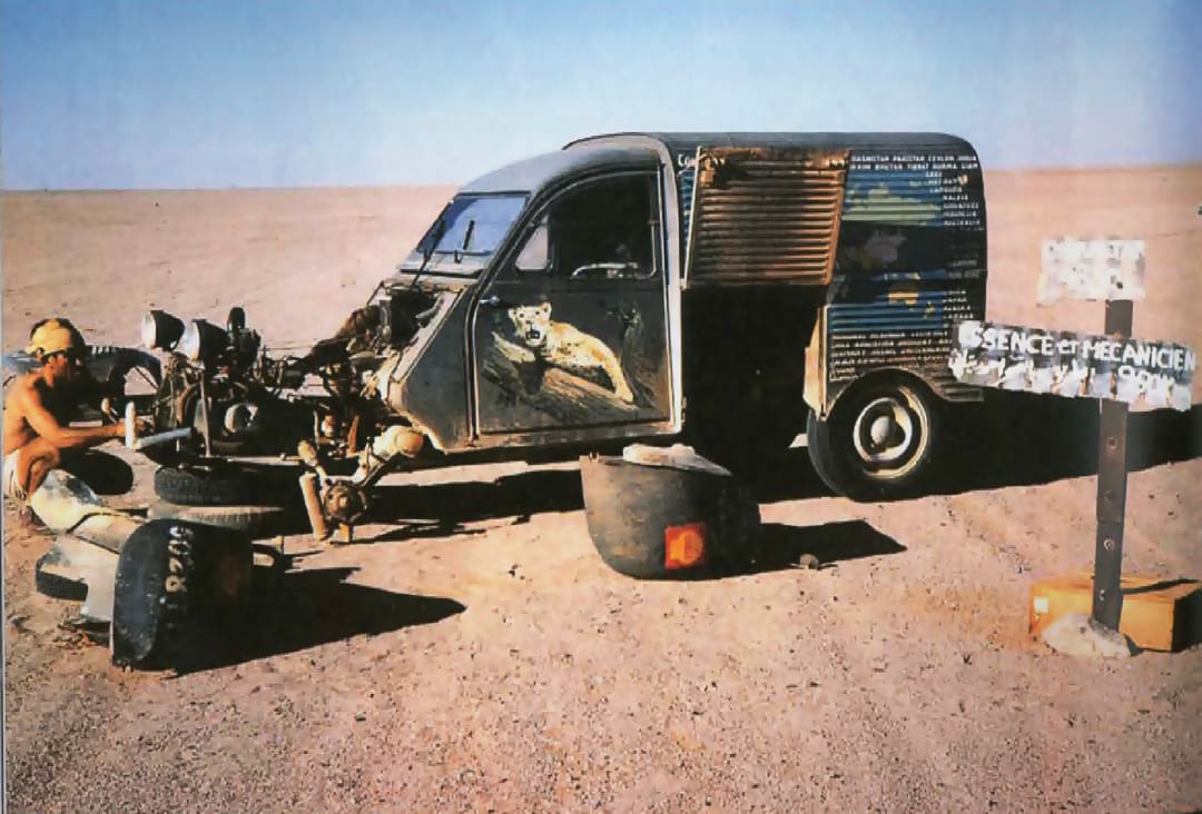 A person repairs a stripped-down car in a barren desert, reminiscent of the Omidvar brothers’ adventures. Animal artwork decorates the side, tires are scattered, and a sign reads SCIENCE ET MECHANIQUE.
