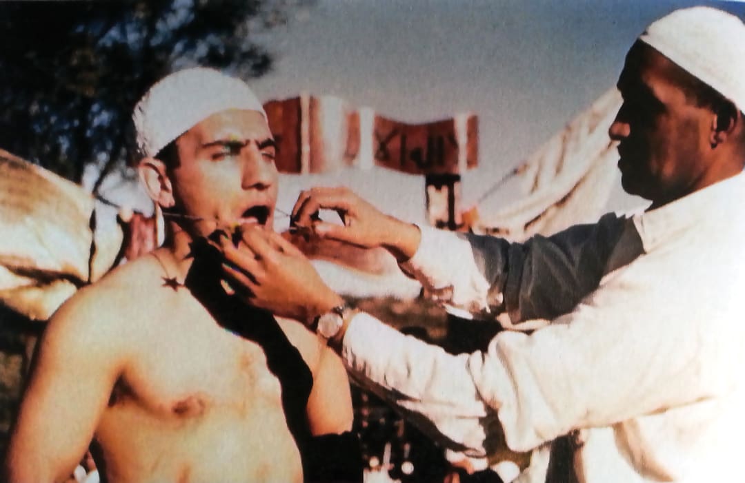A man in a white headwrap stands shirtless as another man, also in a headwrap, appears to insert a sharp object into his mouth during an outdoor ritual, reminiscent of the omidvar brothers’ adventurous journeys.