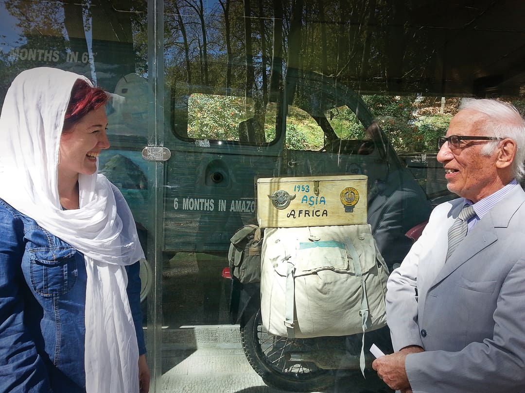 A smiling woman in a white headscarf talks with an older man in a suit near a bicycle with a 1953 Asia Africa sign—reminiscent of the Omidvar brothers—standing before a glass case and vintage vehicle.