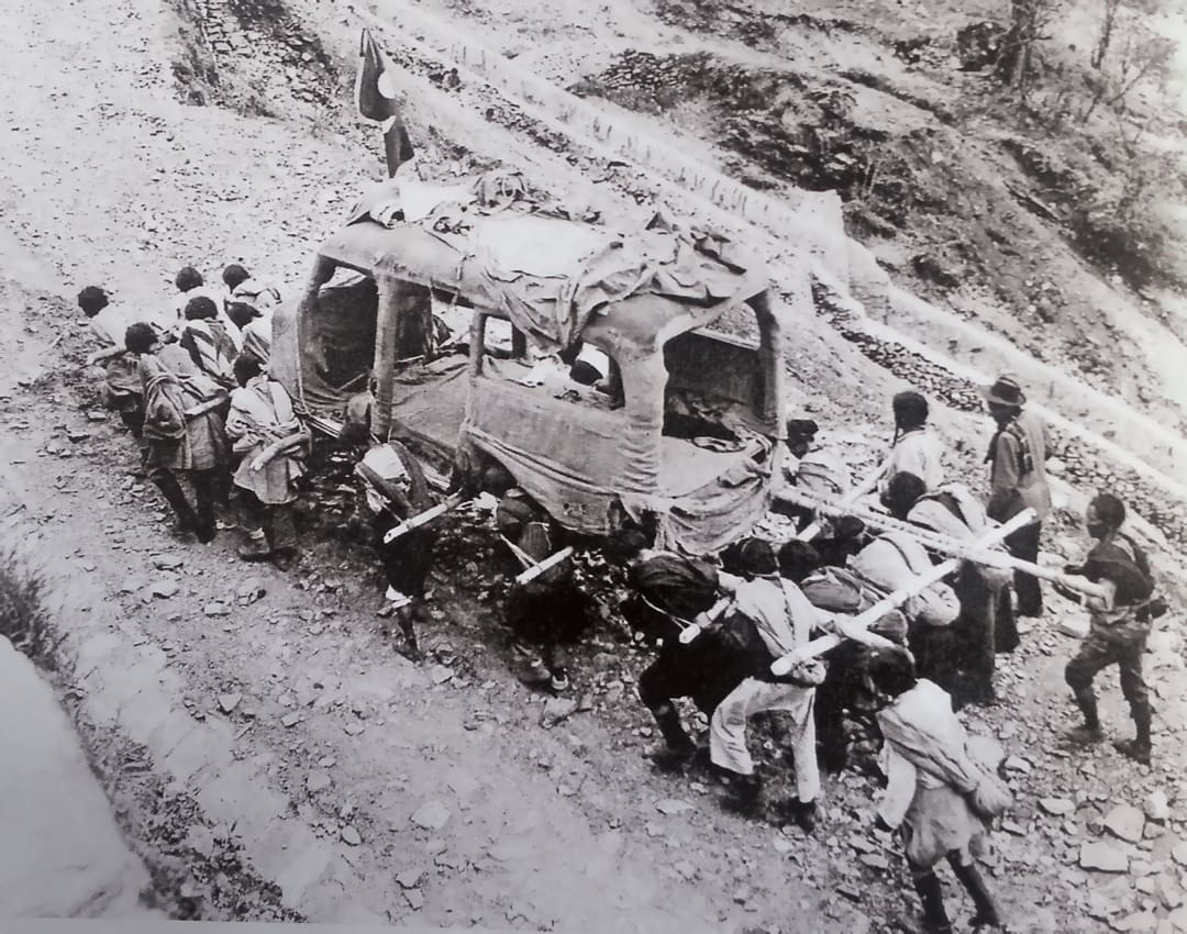 A group of people carry a large, makeshift sedan chair with passengers along a rocky, uphill path in a rural, mountainous area, reminiscent of the adventurous journeys of the Omidvar brothers.