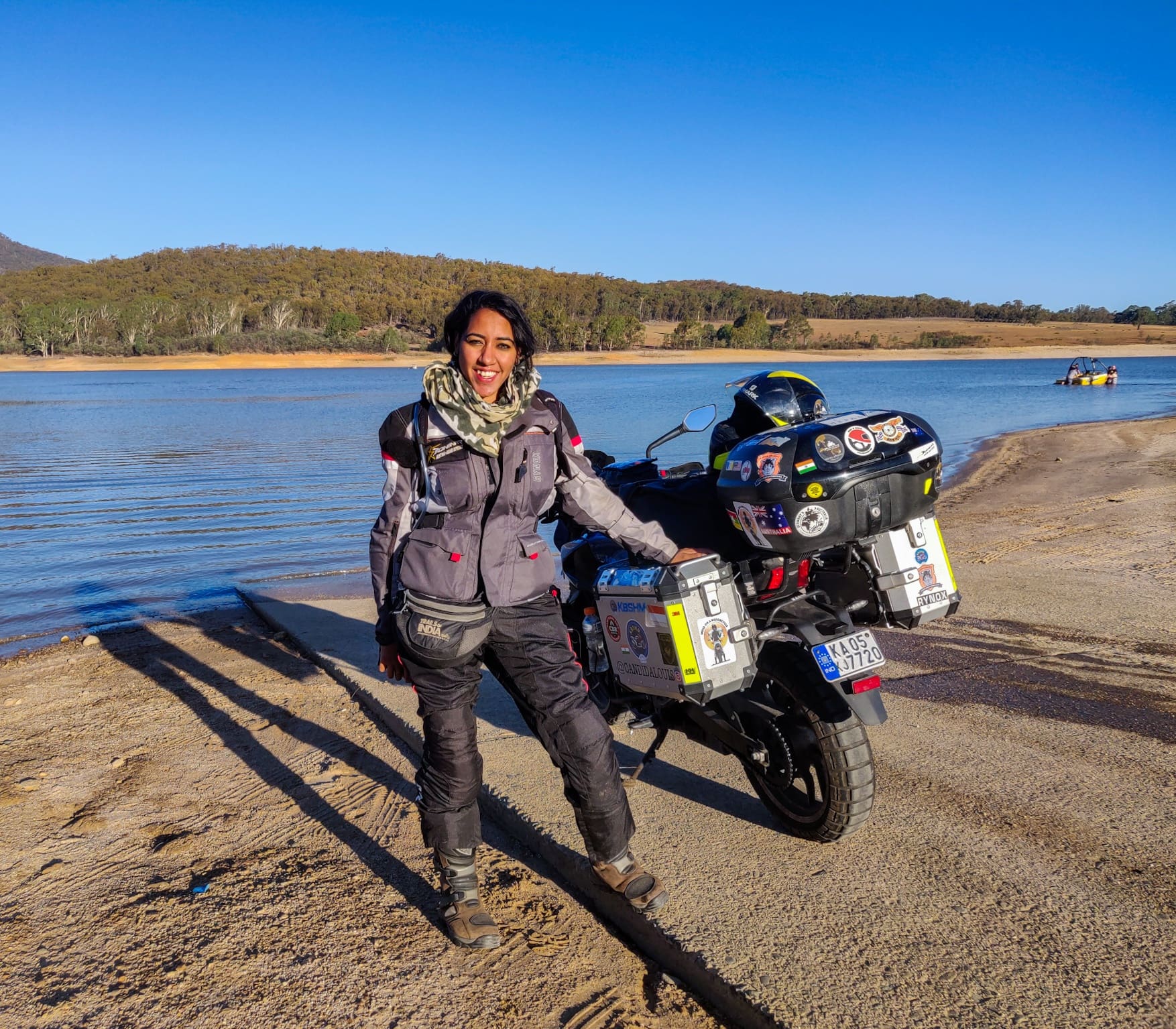 Candida Louis, in full motorcycle gear, stands on a lakeshore smiling, one hand resting on a touring bike covered in travel stickers, with water, forests, and blue sky in the background.