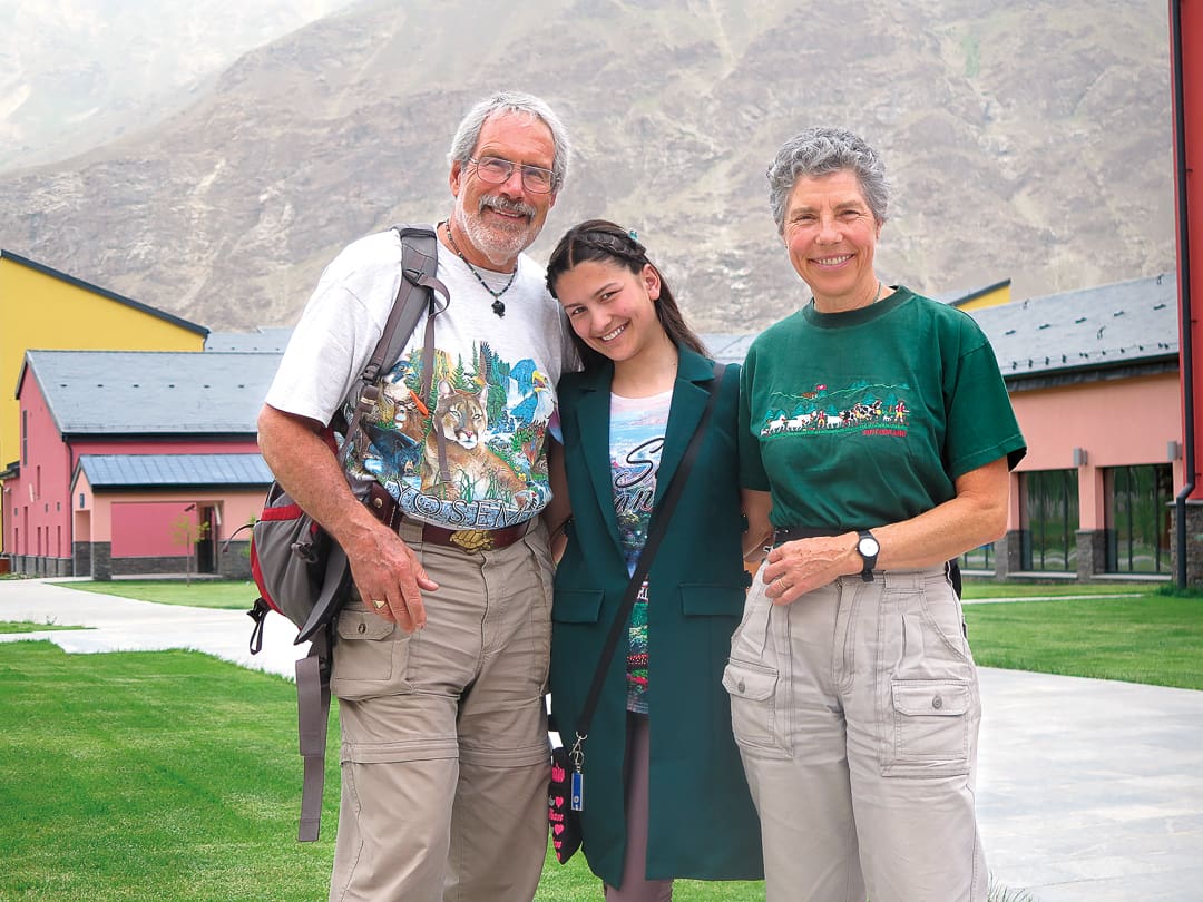 Three people stand smiling outdoors in front of colorful buildings and the majestic Pamirs, with green grass underfoot. Two older adults wear outdoor clothing, and a younger woman stands casually between them.