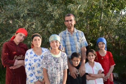 A family of seven stands outdoors amid the lush greenery of the Pamirs, wearing colorful clothing and headscarves. They are posing for the camera and smiling gently in natural light.