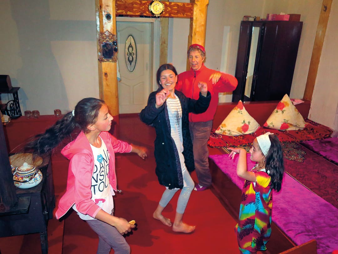 Four people, three children and one adult, dance and smile together in a cozy, warmly lit room with cushions and a carpeted floor—sharing joyful moments reminiscent of family gatherings in the Pamirs.