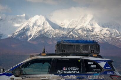 A Subaru rally car equipped with a Leitner Designs roof-mounted storage box parked in front of snow-capped mountains.