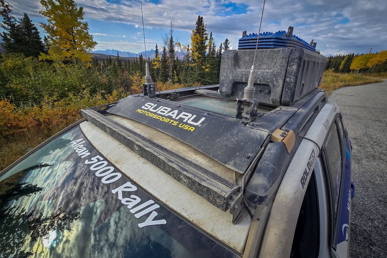 Roof of a Subaru Motorsports USA rally car featuring a Leitner Designs storage container, light bar, and antennas, parked on a road with autumn foliage and mountains in the background.