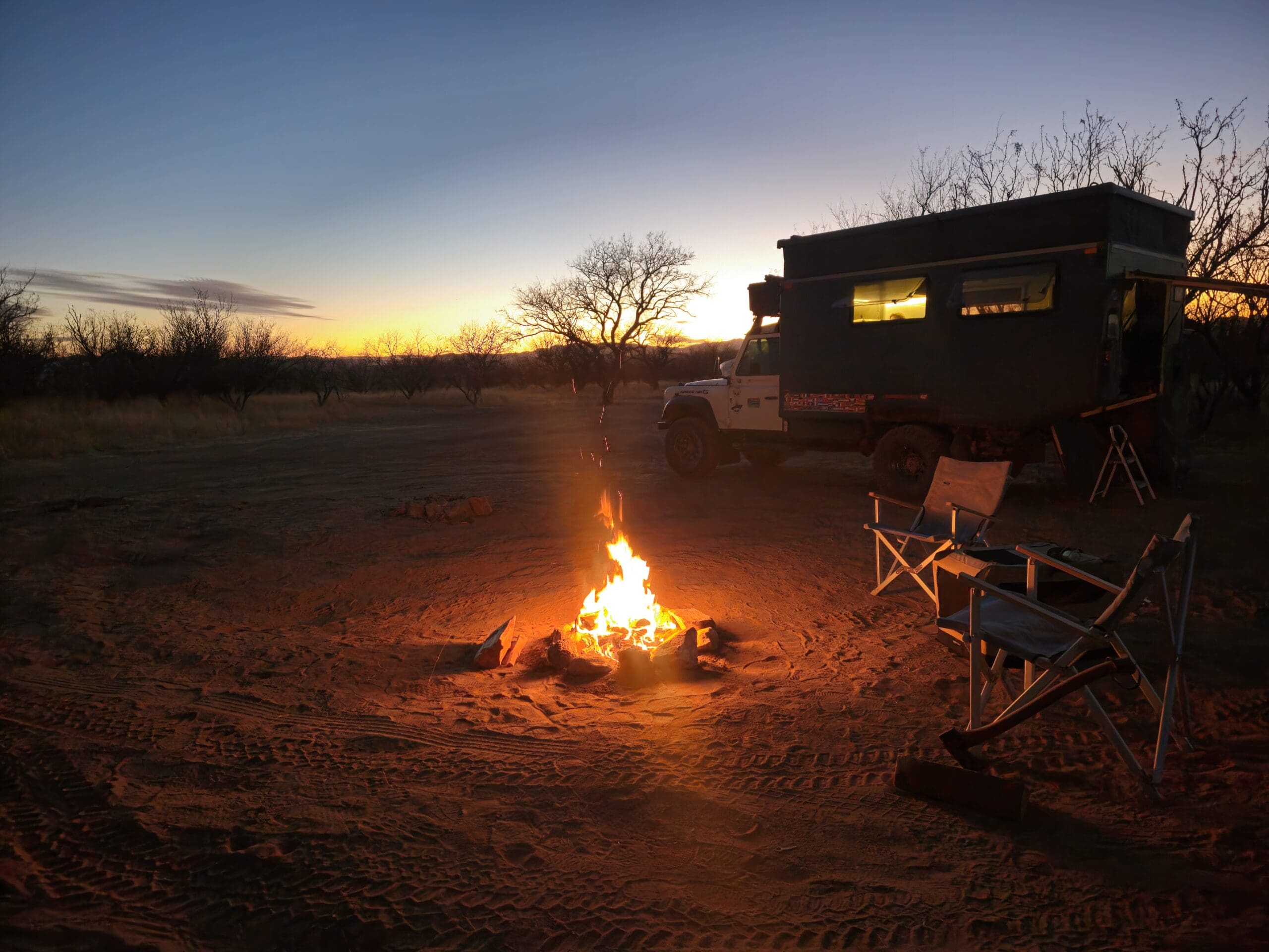 A campfire burns in the sand at sunset next to a parked camper vehicle and two folding chairs along the Arizona Traverse.