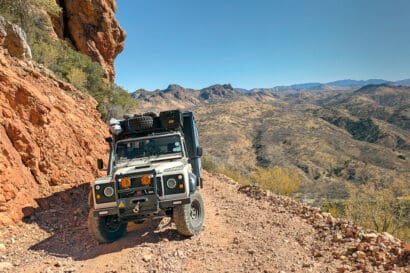 A Land Rover Defender with a roof rack parked on a narrow, rocky mountain trail of the Arizona Traverse, overlooking a vast desert landscape.