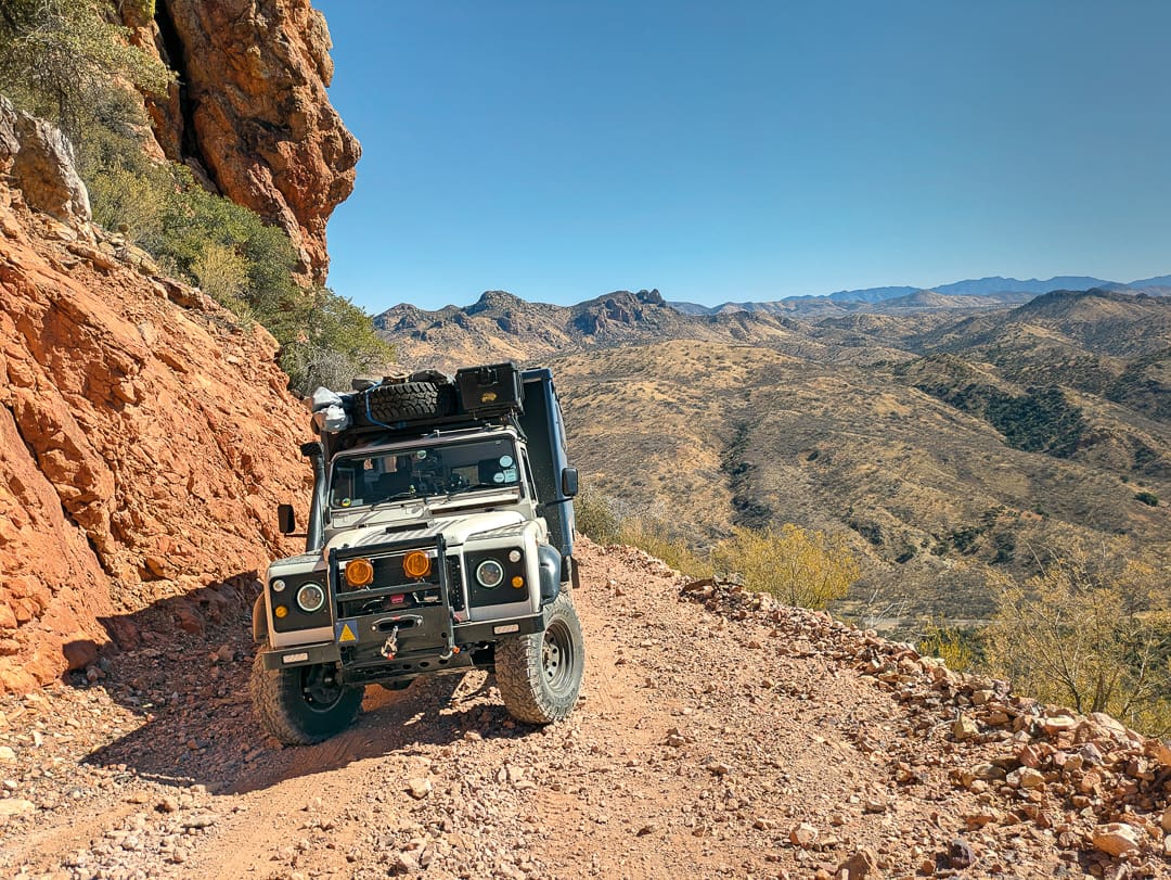 A Land Rover Defender with a roof rack parked on a narrow, rocky mountain trail of the Arizona Traverse, overlooking a vast desert landscape.