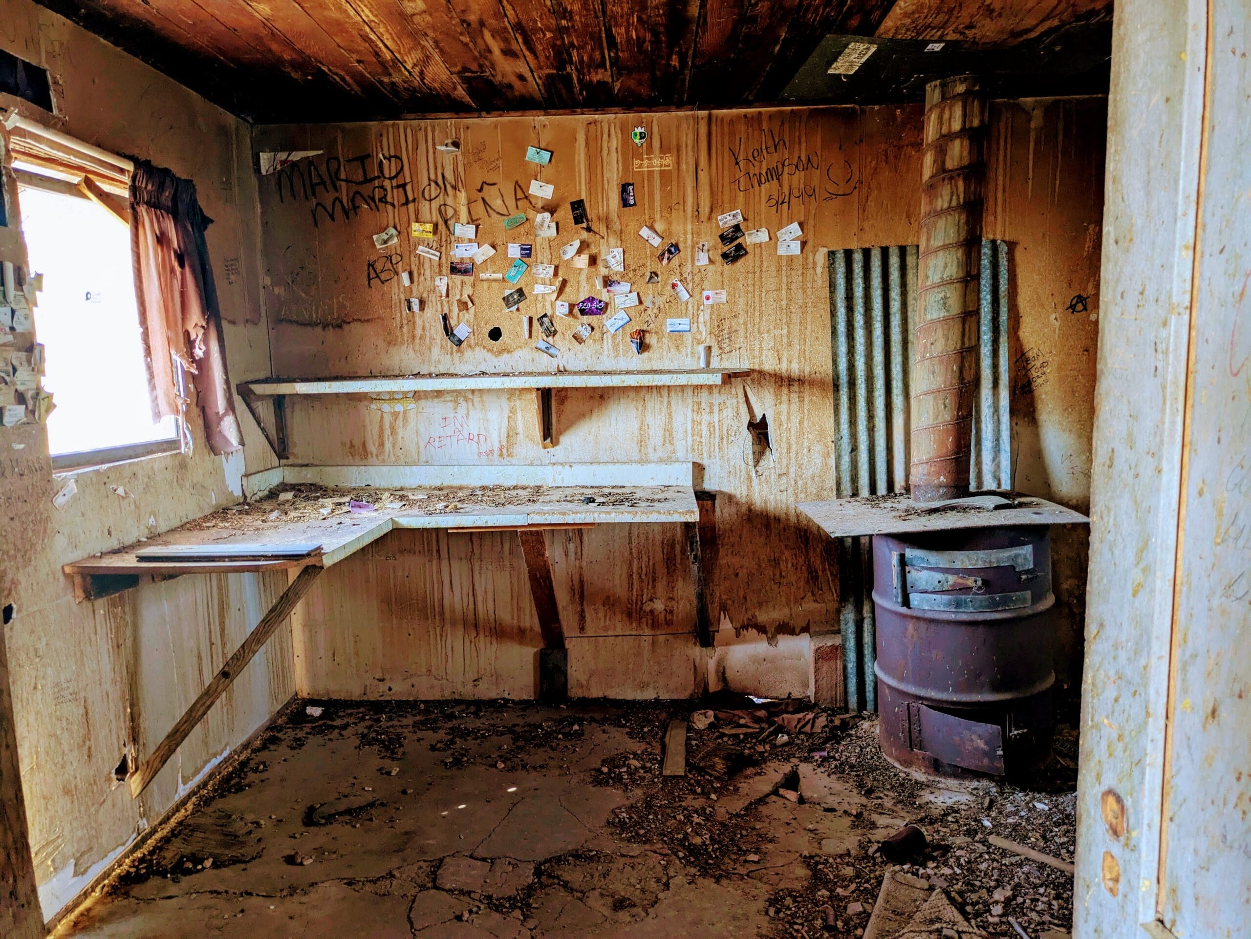 A dilapidated room with graffiti on the walls, a collection of OV Routes business cards pinned to the wood, a workbench, and a rusted metal stove.