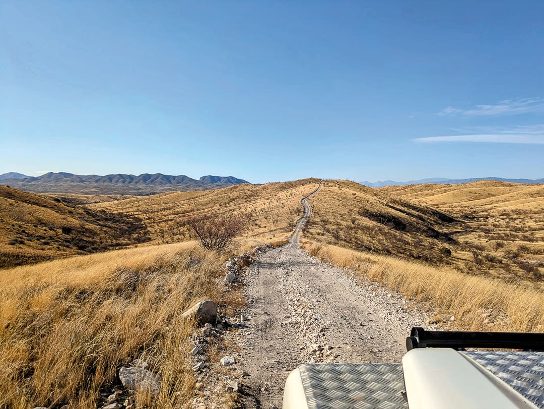 A gravel road on the Arizona Traverse stretches over dry, rolling hills under a clear blue sky, as seen from the hood of a vehicle.