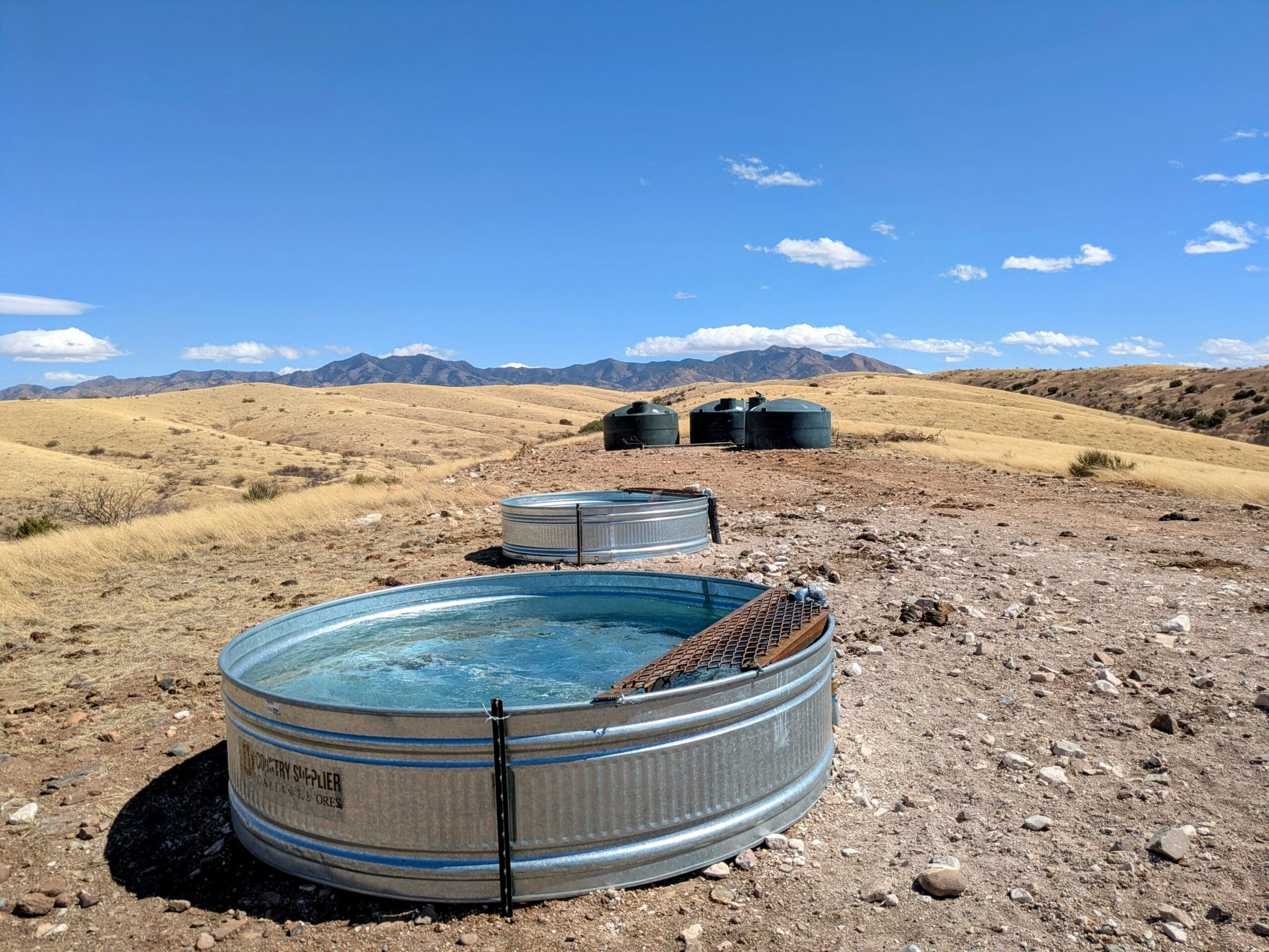 Two round metal stock tanks filled with water and three dark plastic water storage tanks set in a dry, mountainous landscape along the Arizona Traverse.