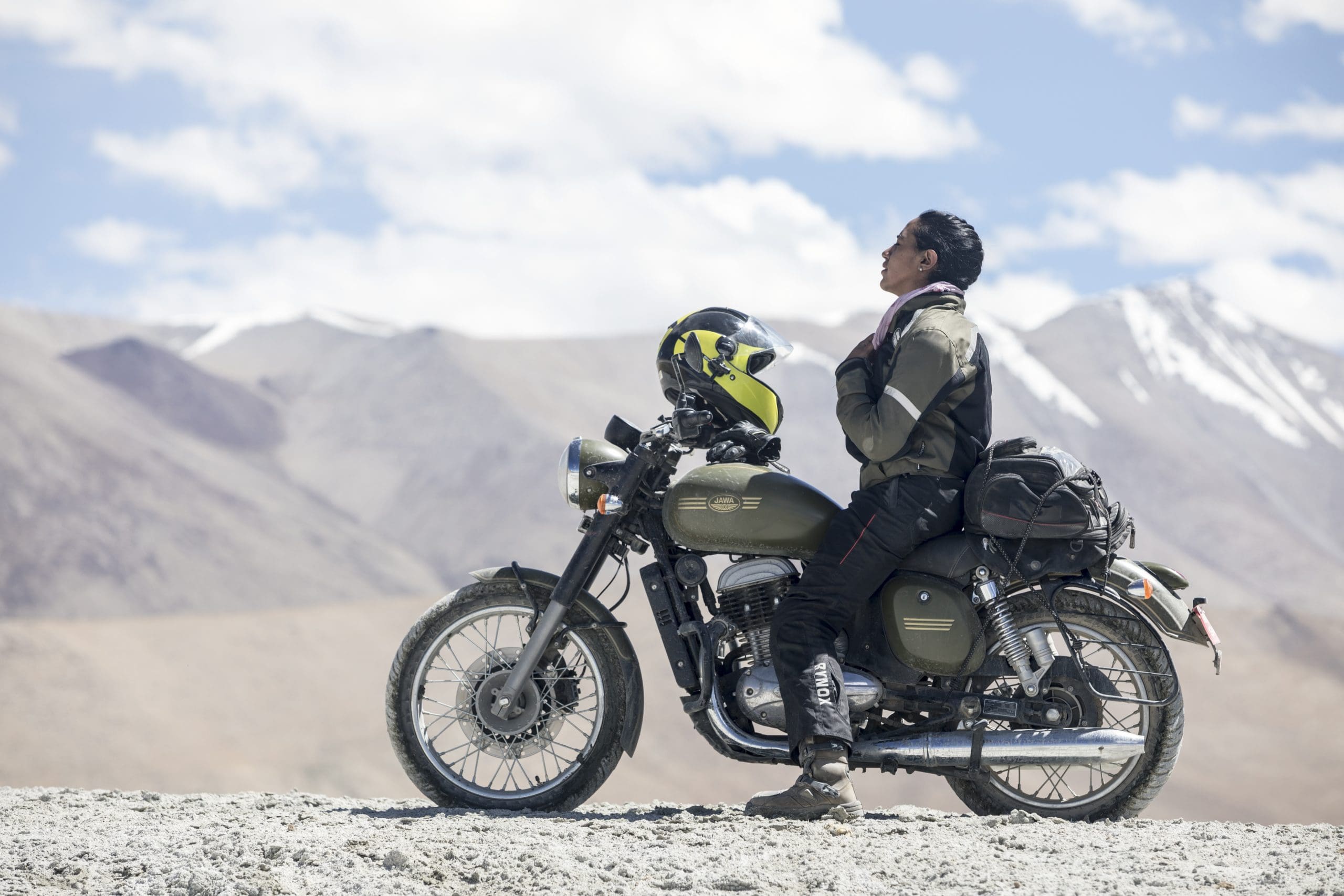 Candida Louis motorcycle gear stands beside an olive-green motorcycle, gazing upward with snow-capped mountains and a bright sky behind—an adventure reminiscent of Candida Louis. A helmet rests on the bike’s seat.
