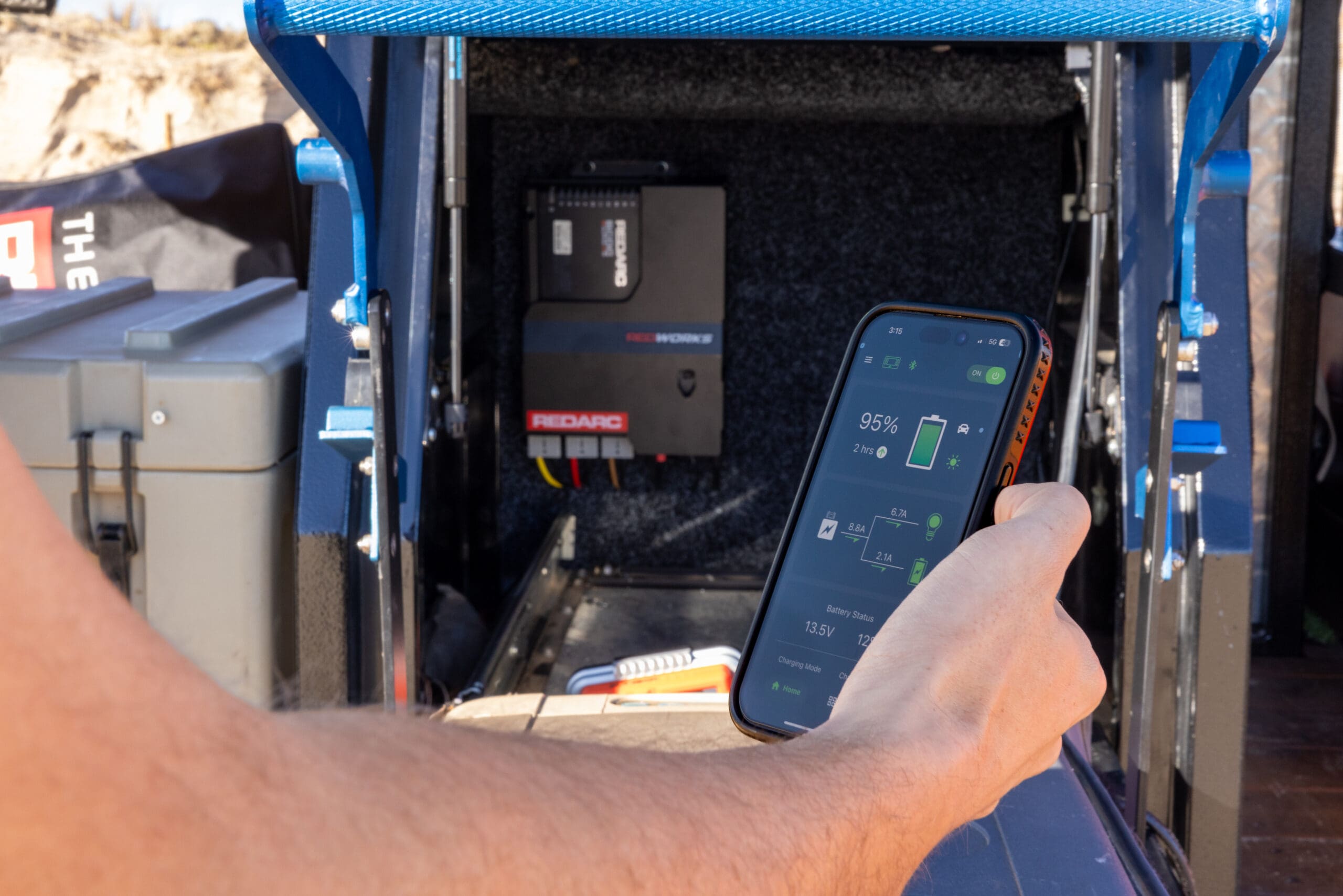 A hand holds a smartphone displaying a 95% charge level in a battery management app, showing the latest overland updates in front of a Redarc battery system installed in a vehicle storage compartment.