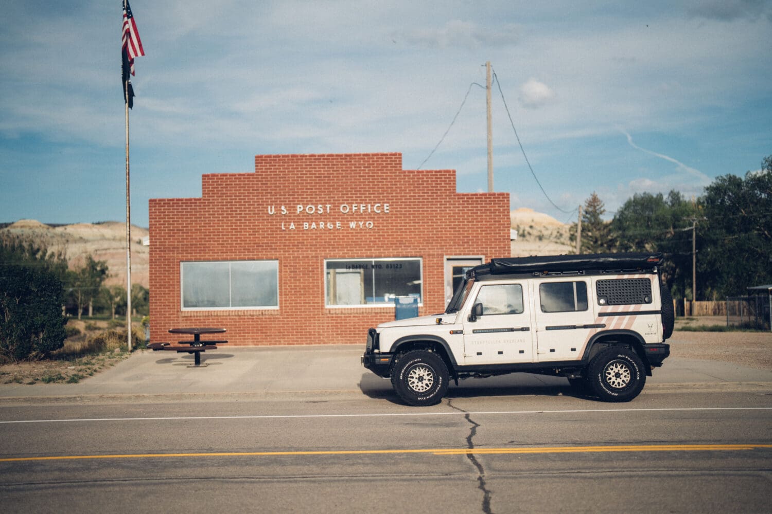 A white SUV is parked in front of a small red brick U.S. Post Office in La Barge, Wyoming, reminiscent of scenes in Cooke City. An American flag flies on a pole nearby.