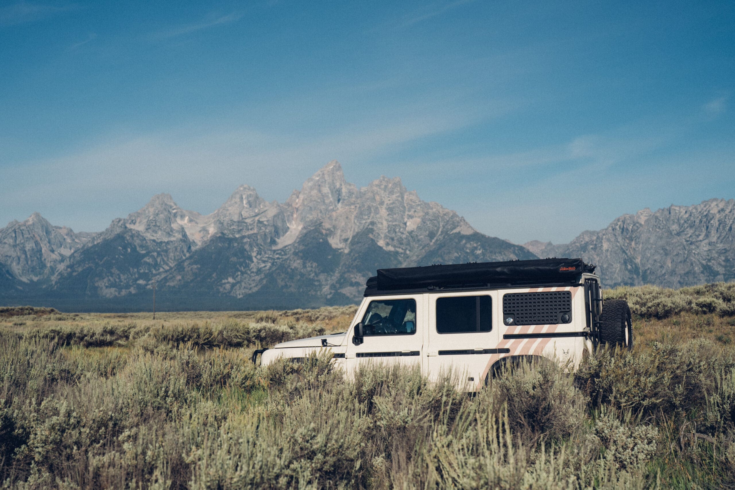 A white SUV with rooftop cargo is parked in a grassy field near Cooke City, with tall mountains and a blue sky in the background.
