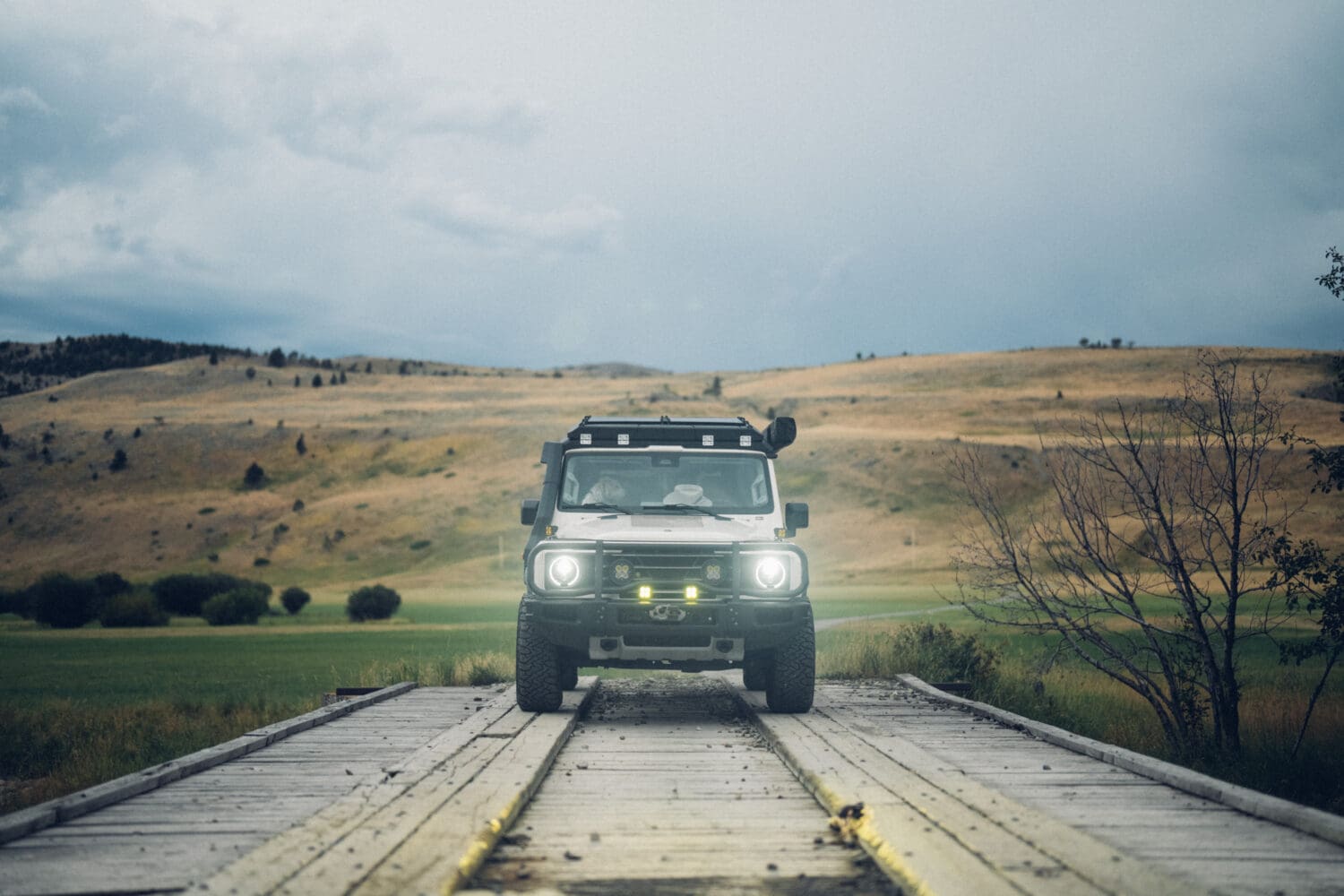 A white off-road vehicle with headlights on drives across a wooden bridge near Cooke City, set against a rural landscape of rolling hills and cloudy skies.