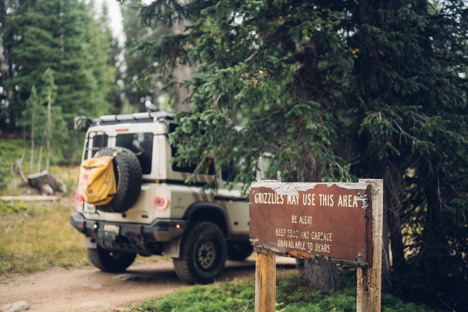 A jeep with a yellow backpack is parked on a dirt trail near Cooke City’s forest. A wooden sign in the foreground warns that grizzly bears may use the area and advises visitors to stay alert.