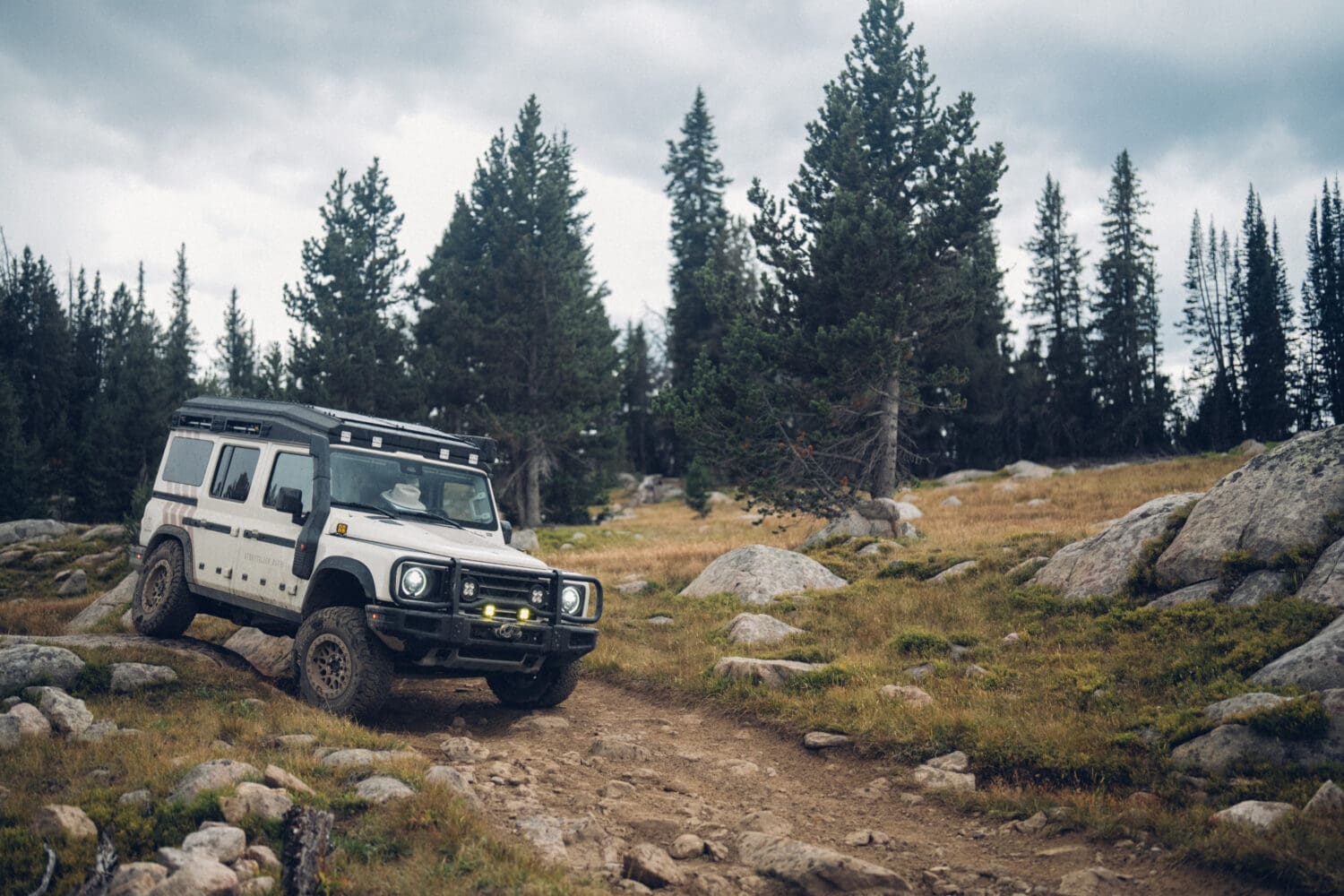 A white off-road vehicle drives on a rocky dirt trail through a pine forest near Cooke City, surrounded by grass, rocks, and cloudy skies, suggesting a rugged outdoor adventure.