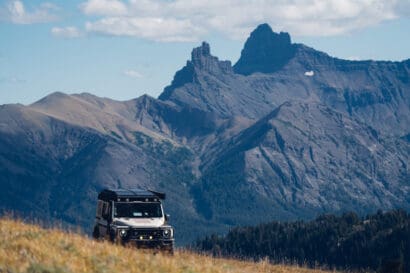 A rugged off-road vehicle drives on grassy terrain near Cooke City, with dramatic, rocky mountains and a few clouds in the background under a blue sky.
