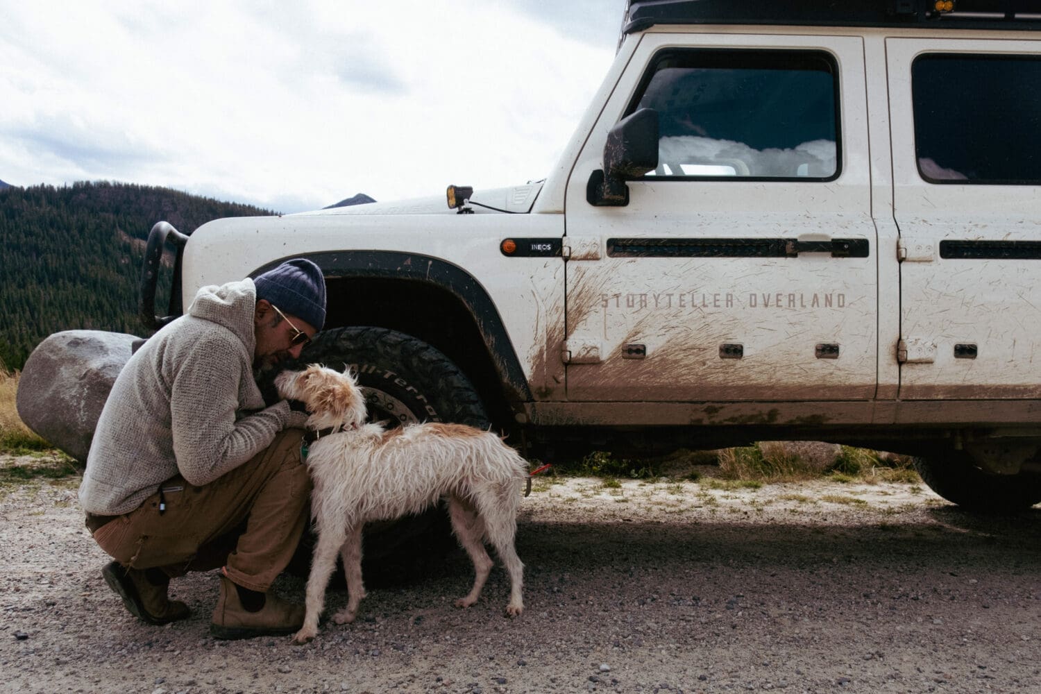 A person in a beanie kneels beside a muddy off-road vehicle near Cooke City, gently touching noses with a wet, white dog. Mountains and trees are visible in the background.