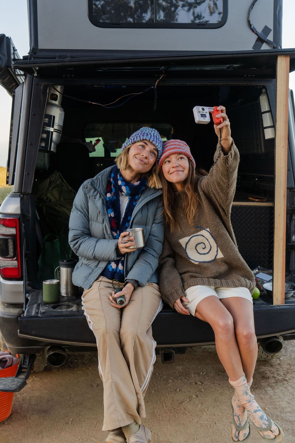 Two women in beanies sit on a lowered truck tailgate, taking a selfie with a small digital camera while capturing overland updates.