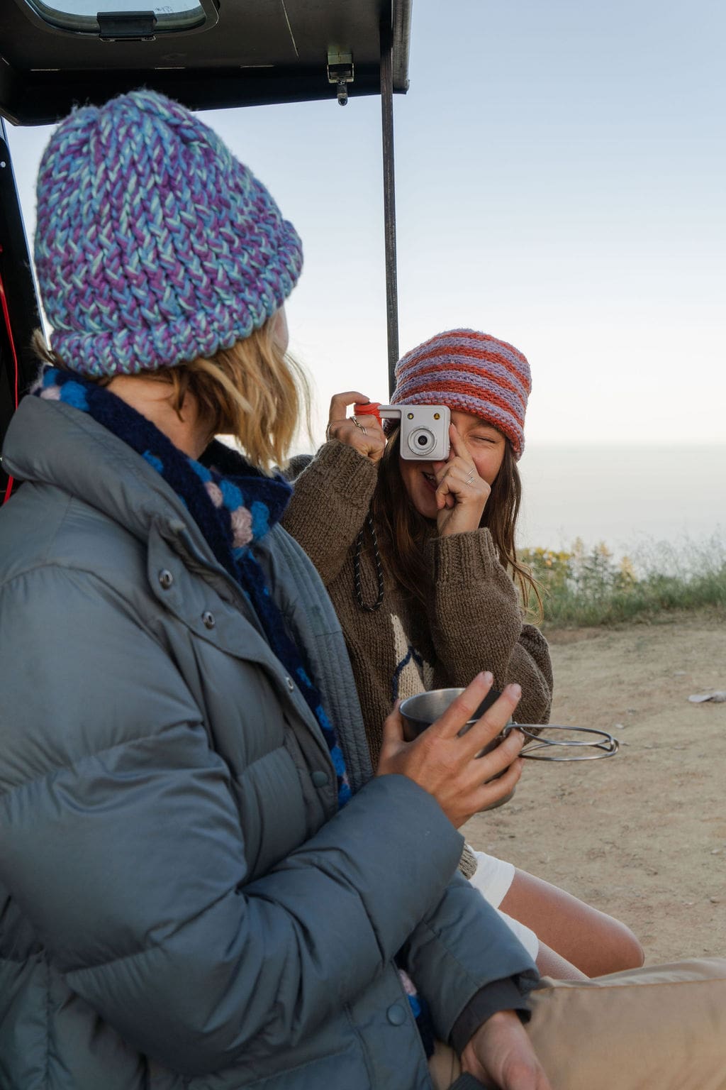 A woman in a striped beanie takes a photo of another woman with a small instant camera during their adventure travel.