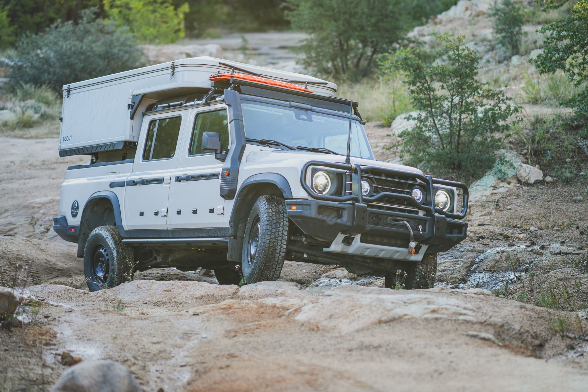 A white XOVERLAND off-road truck with a camper shell driving over rocky terrain.