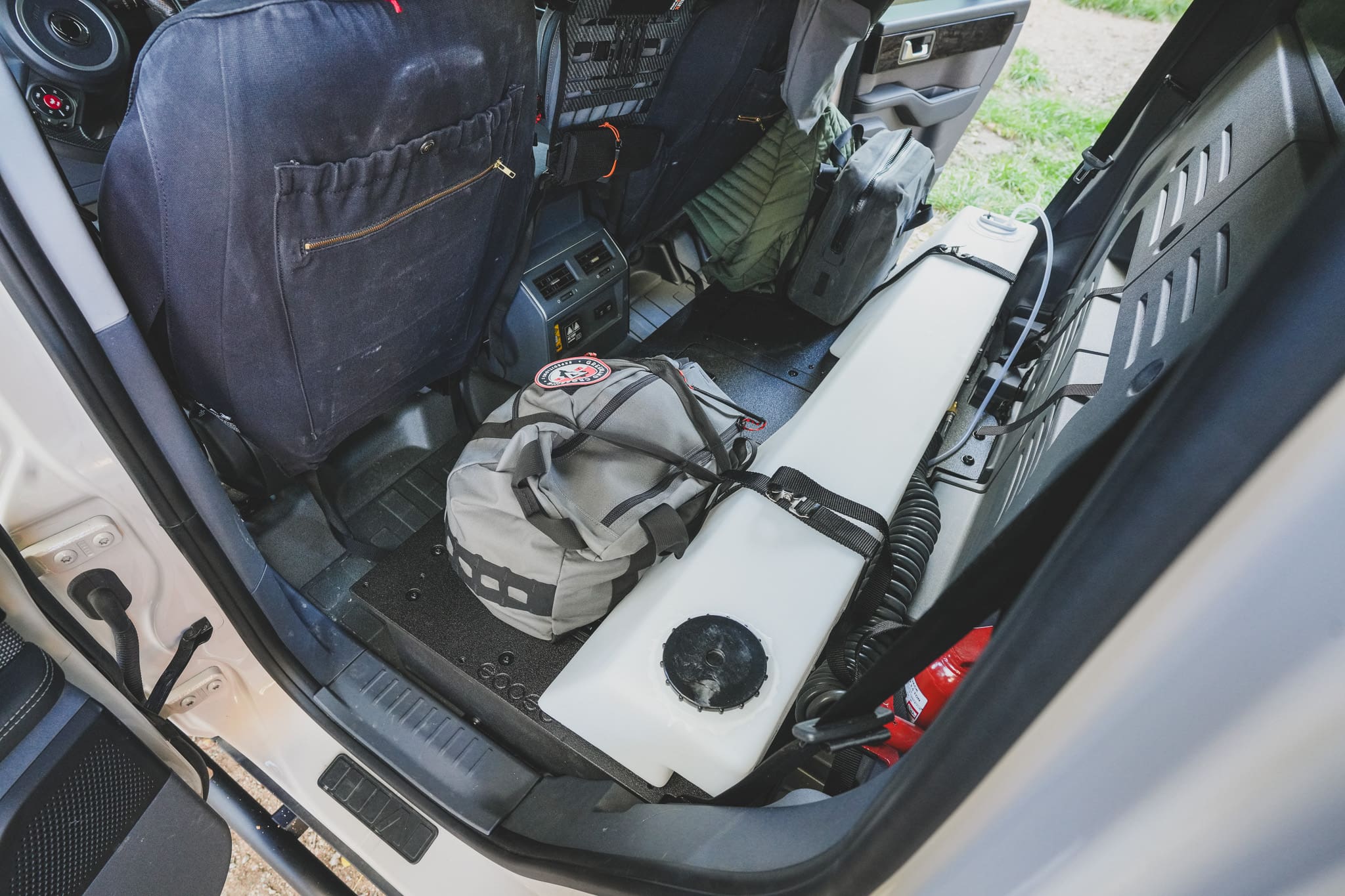 Interior view of a vehicle’s back seat area, showing an FHF storage platform with a grey gear bag and a large white water tank as part of the XOVERLAND new drive line gear setup.