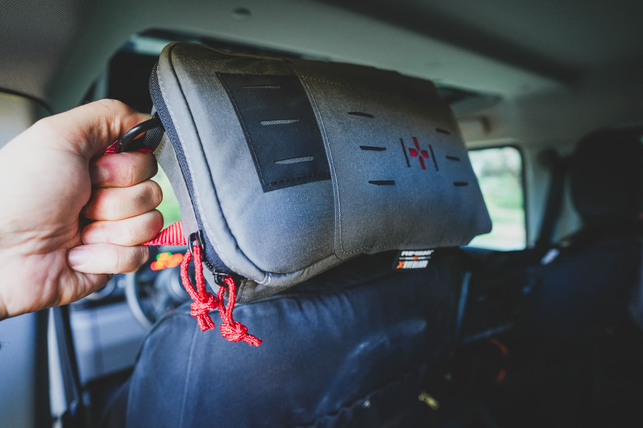 A hand holding a gray XOVERLAND tactical medical kit with a red cross emblem, hanging from the back of a car seat headrest.