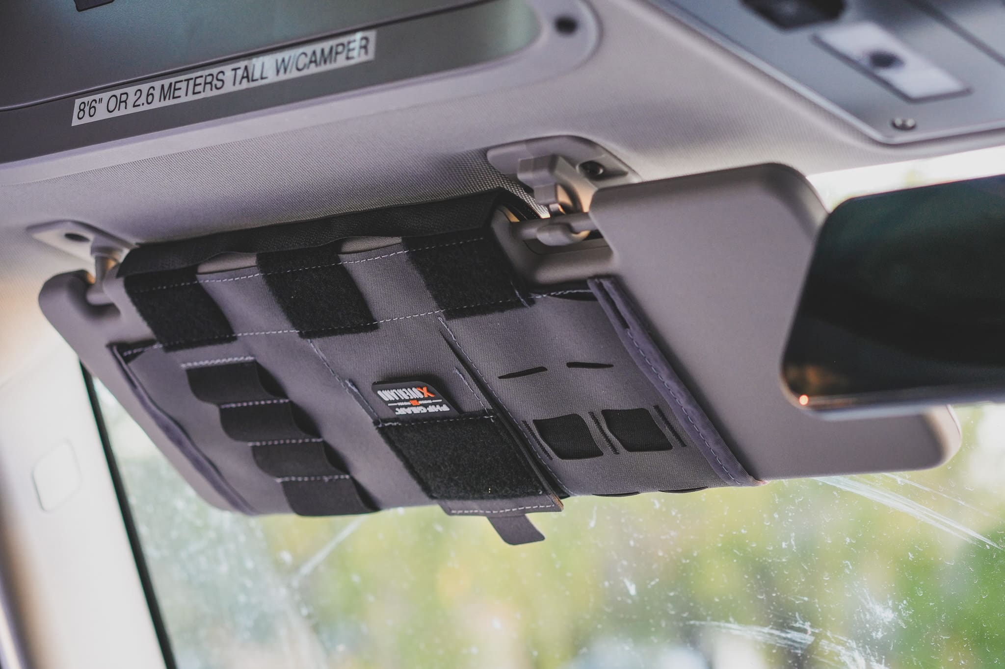 A gray XOVERLAND modular visor organizer with MOLLE webbing and an FHF logo patch attached to a vehicle sun visor, with a height clearance sticker visible on the ceiling above.