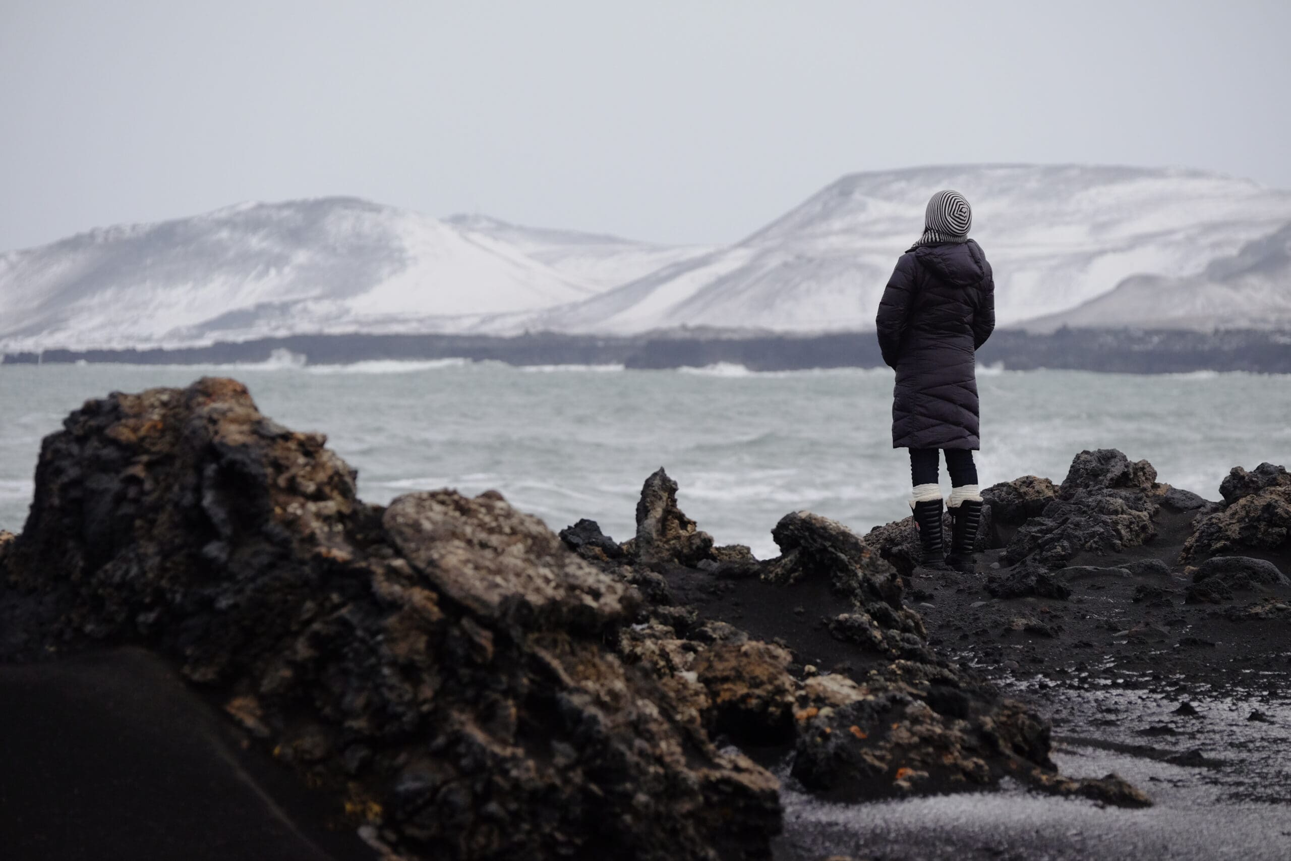 Stephanie Brady stands on a rocky black beach in a winter coat and hat, looking out at a turbulent sea and snow-covered mountains.